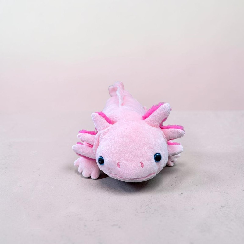 Ein flauschiges, pinkes Axolotl-Plüschtier liegt auf einer hellgrauen Oberfläche. Es hat große, buschige pinke Kiemen, kleine schwarze Kulleraugen und ein leichtes Lächeln. Der Hintergrund ist sanft rosa und unscharf.
